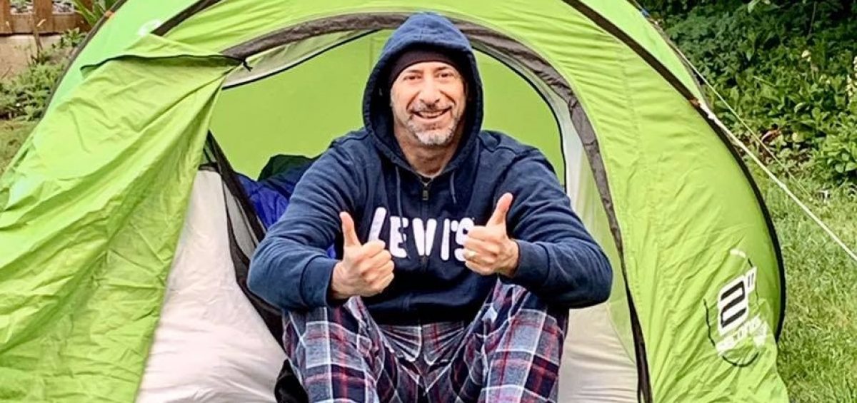 man sat in green tent with thumbs up, wearing a blue hooded top and checked trousers