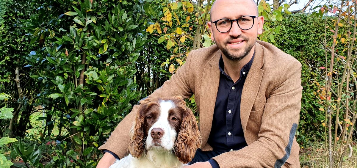 man in beige jacket and blue jeans, kneeling down, with a springer spaniel dog at his side. In the background are green leaf trees